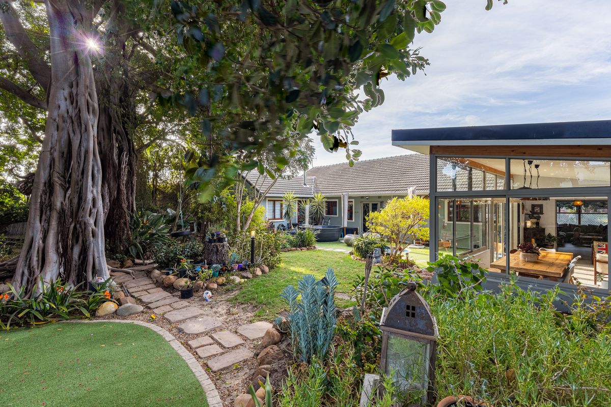 A well-maintained rear garden featuring a mature tree, stone pathway, artificial lawn, and a modern glass-enclosed patio extension.