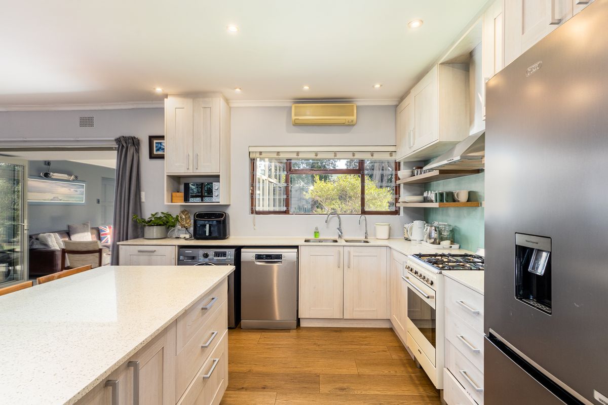 Modern kitchen featuring light wood cabinetry, stone countertops, a central island, and integrated appliances, with an open-plan flow to the adjacent living area.