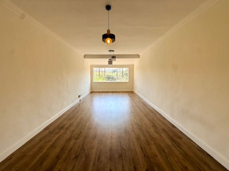 A spacious, empty living area featuring laminate flooring, coved ceilings, and modern pendant lighting, illuminated by a large window.