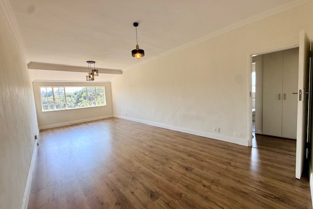 A bright, empty living room featuring laminate flooring, modern pendant lighting, and coving, with a view through the window and access to a bedroom.