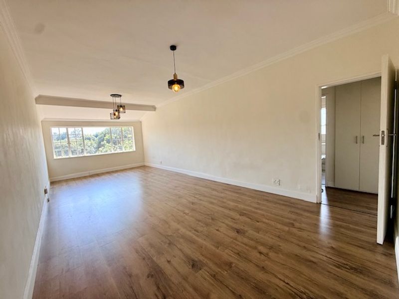 A bright, empty living room featuring laminate flooring, modern pendant lighting, and coving, with a view through the window and access to a bedroom.