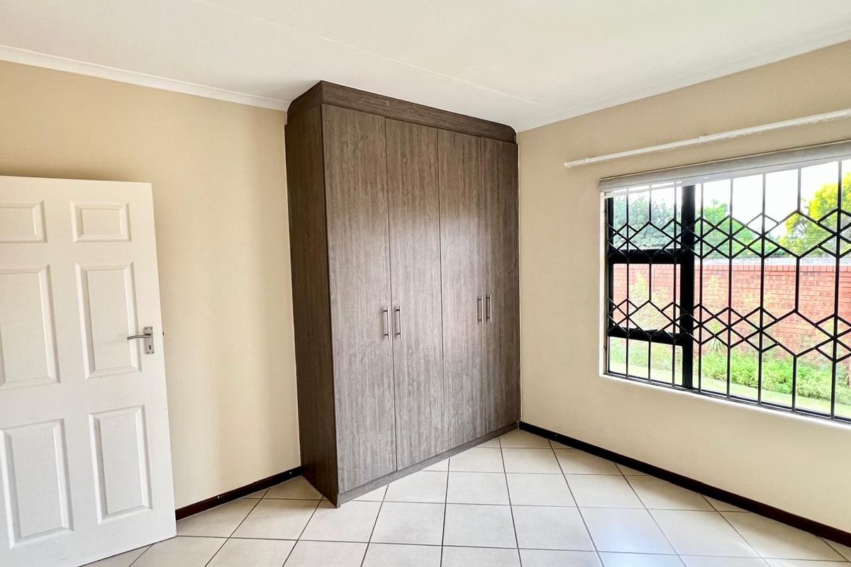 A bright, empty bedroom featuring neutral tiled flooring, a large built-in wooden wardrobe, and windows fitted with security bars.