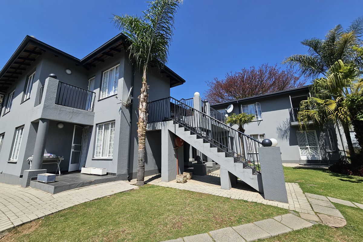 Rear view of a multi-story apartment complex featuring grey exterior walls, an external staircase with iron railings, and a landscaped garden area.