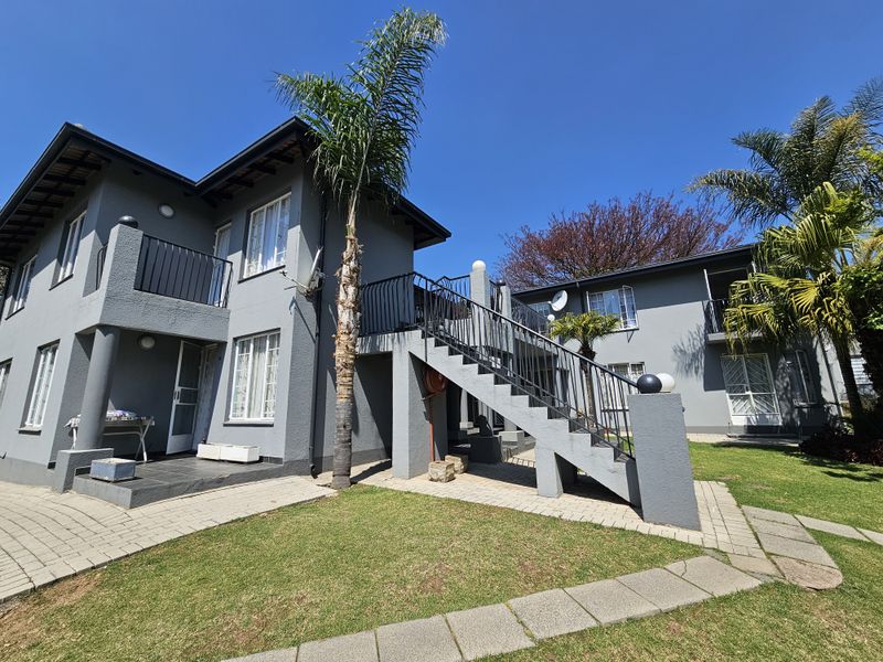 Rear view of a multi-story apartment complex featuring grey exterior walls, an external staircase with iron railings, and a landscaped garden area.