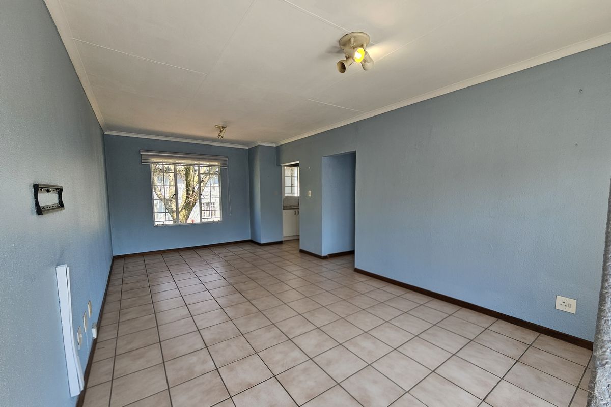 A spacious, empty living area featuring neutral tiled flooring, blue painted walls, and natural light from a window with security bars.