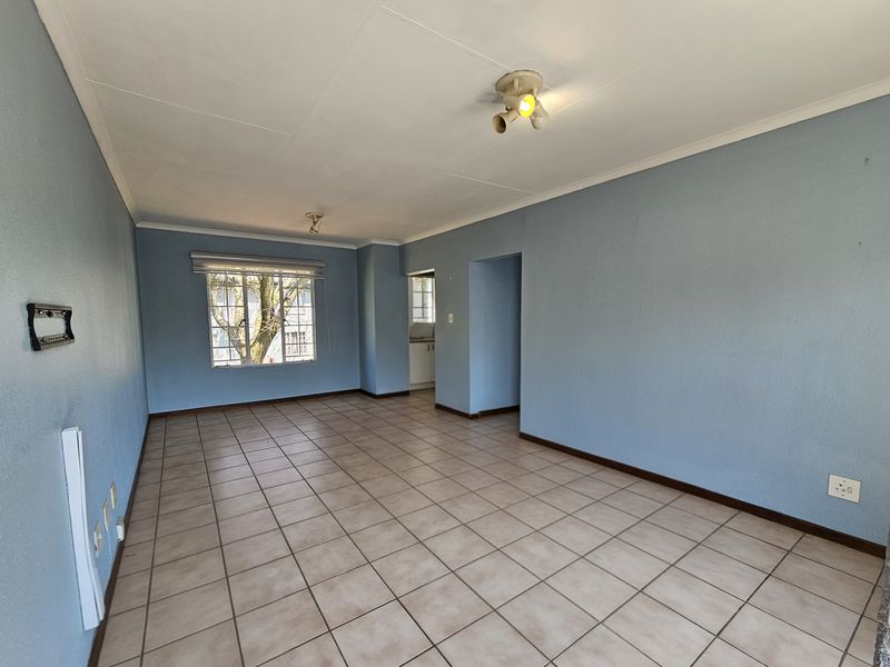 A spacious, empty living area featuring neutral tiled flooring, blue painted walls, and natural light from a window with security bars.