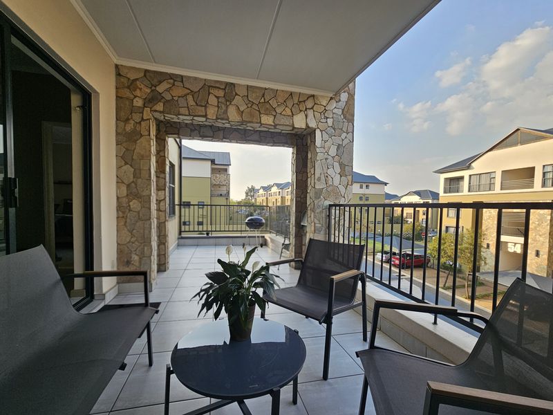 A spacious covered balcony featuring stone cladding accents, tiled flooring, and a metal balustrade, offering views of the surrounding apartment complex.