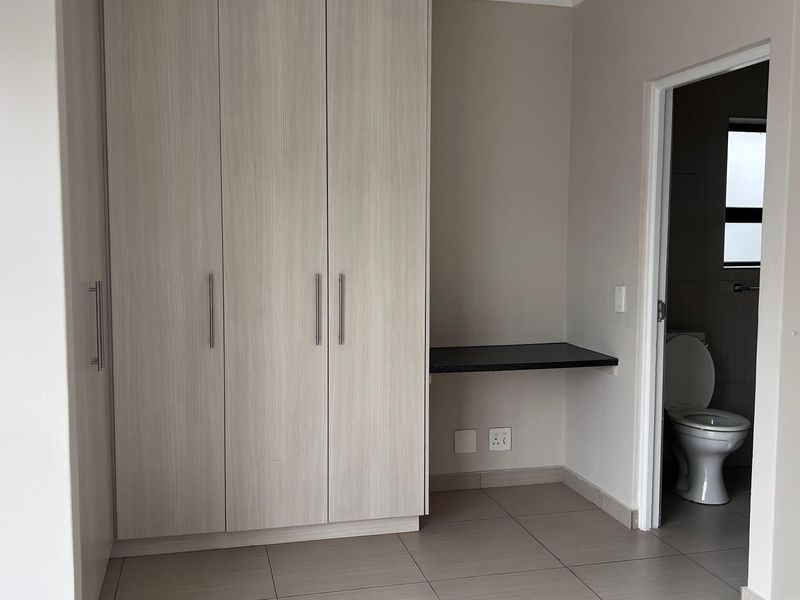 A clean, modern bedroom featuring light wood-grain built-in cupboards, a floating desk, tiled flooring, and direct access to an en-suite bathroom.