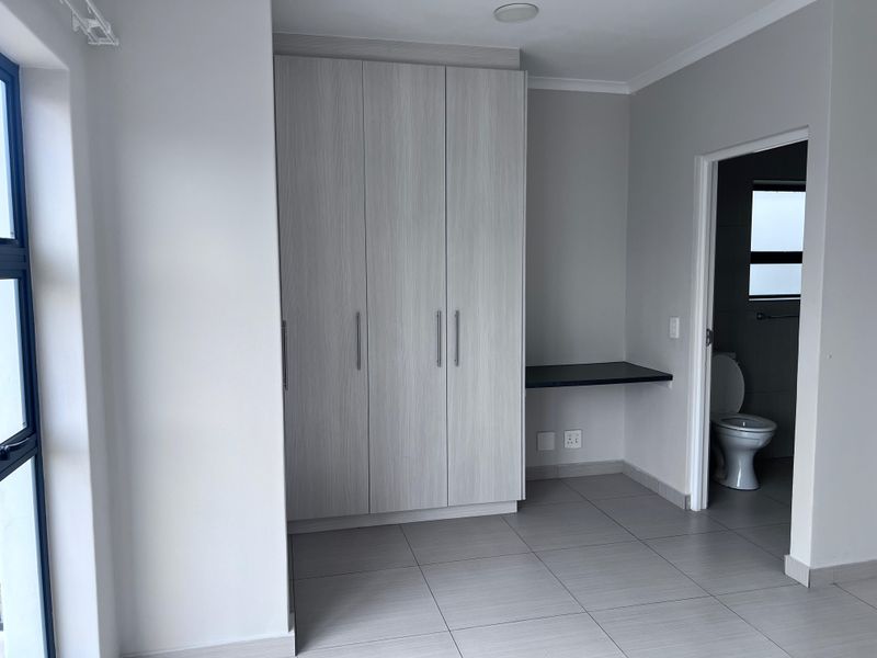 A bright, modern bedroom featuring built-in cupboards, a dedicated desk area, tiled flooring, and direct access to an en-suite bathroom.