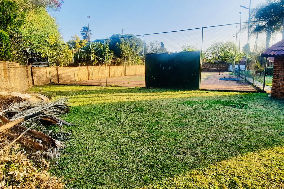 A private residential tennis court enclosed by chain link fencing, featuring an adjacent brick outbuilding and surrounding garden area.