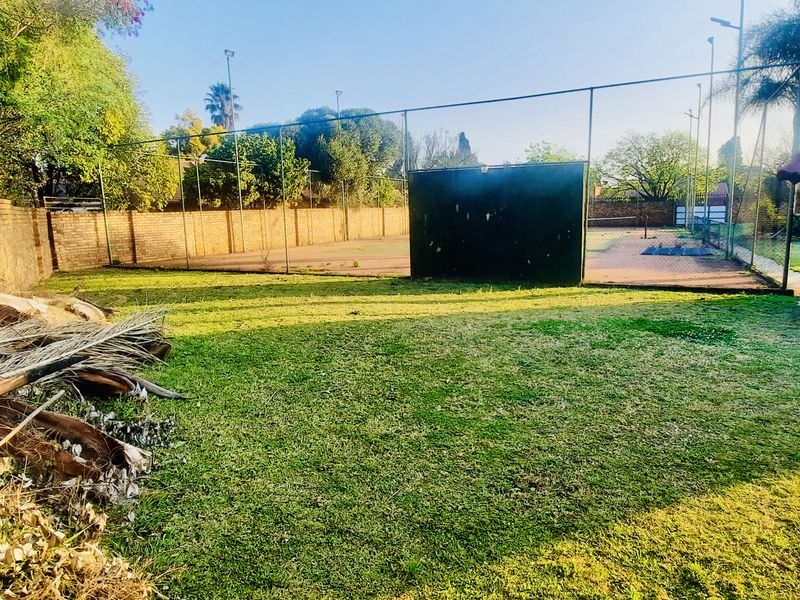 A private residential tennis court enclosed by chain link fencing, featuring an adjacent brick outbuilding and surrounding garden area.