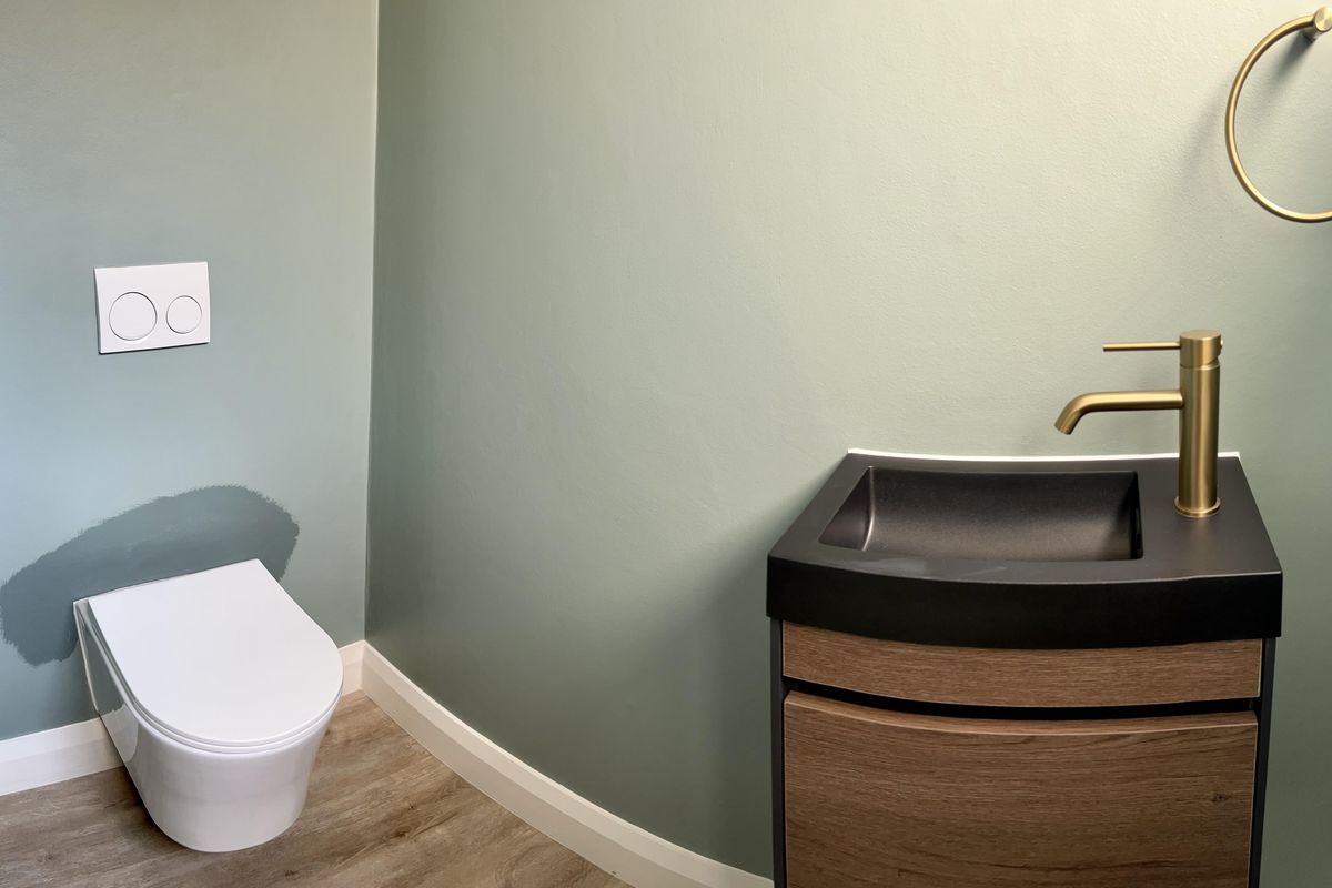 Modern powder room featuring a wall-hung toilet, sleek vanity with integrated basin, and contemporary brass fixtures against sage green walls.