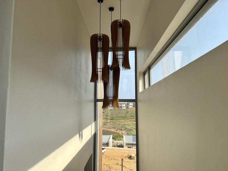 A bright, modern hallway featuring high ceilings, large fixed glass windows, and contemporary pendant lighting with views of the surrounding landscape.
