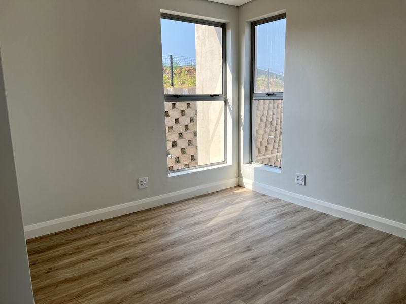 A bright, empty bedroom featuring modern laminate flooring, neutral walls, and corner windows with views of the retaining wall.