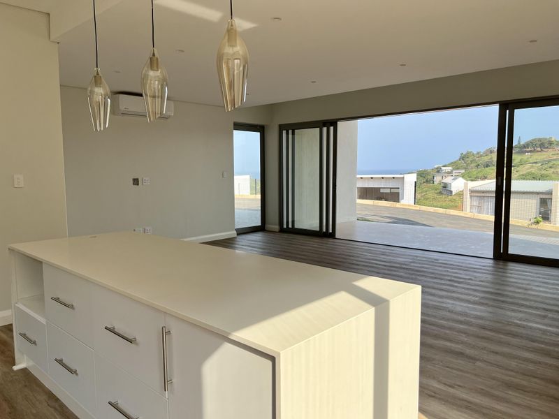 Modern open-plan living area featuring a central kitchen island, sleek laminate flooring, and large sliding glass doors opening to an outdoor patio with coastal views.