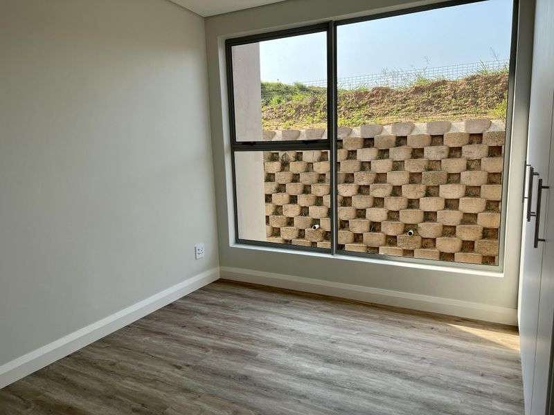 A bright, modern bedroom featuring neutral walls, wood-look laminate flooring, built-in cupboards, and a large window overlooking a landscaped retaining wall.