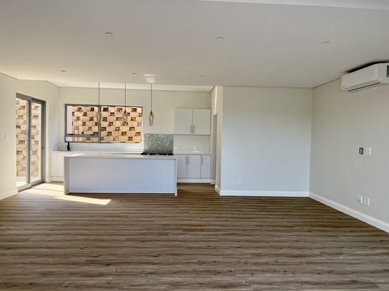 Modern open-plan living and kitchen area featuring sleek white cabinetry, a herringbone backsplash, wood-look laminate flooring, and a wall-mounted air conditioning unit.