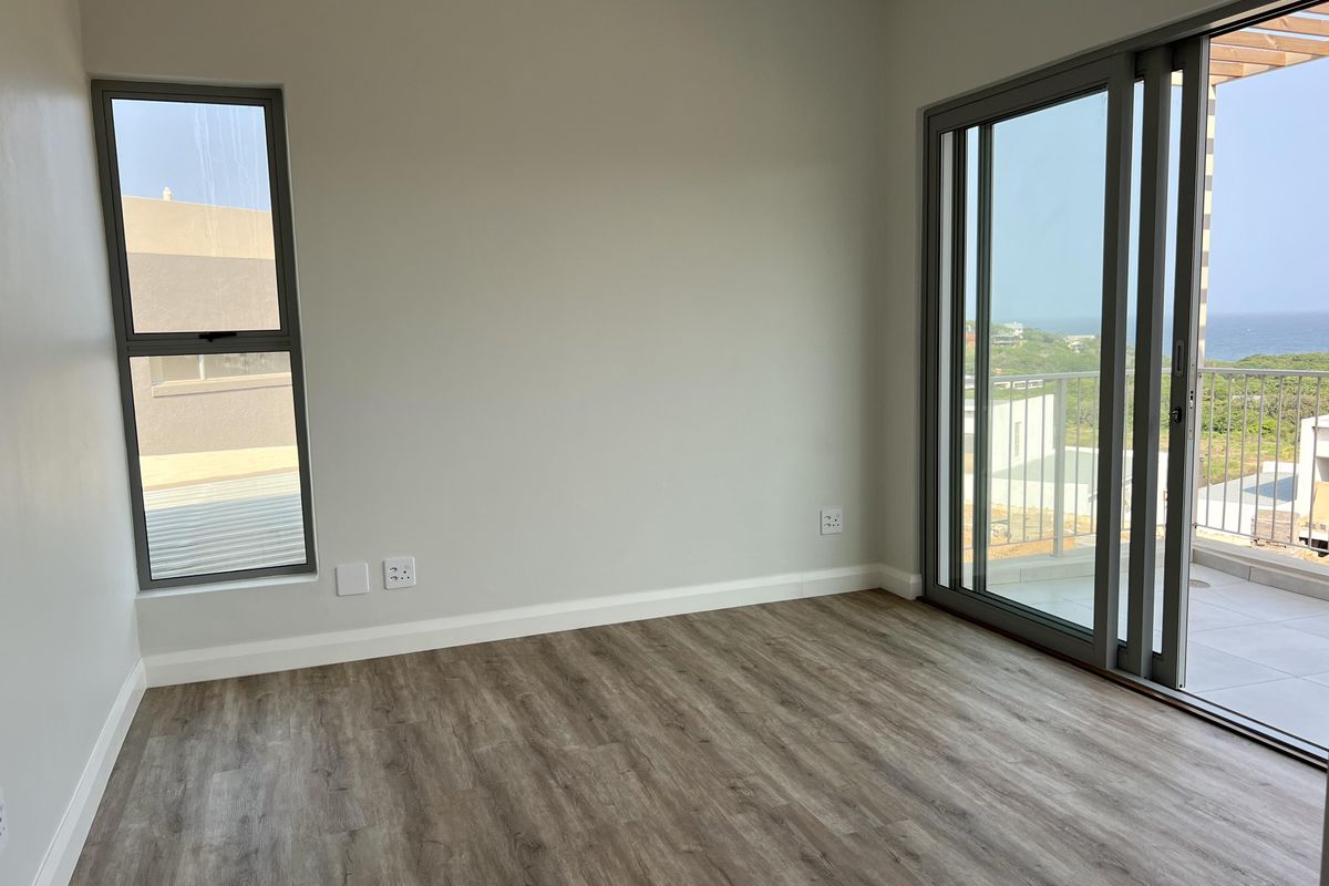 A bright, empty bedroom featuring modern laminate flooring, neutral walls, and large aluminium sliding doors opening onto a balcony with coastal views.