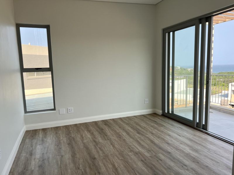 A bright, empty bedroom featuring modern laminate flooring, neutral walls, and large aluminium sliding doors opening onto a balcony with coastal views.