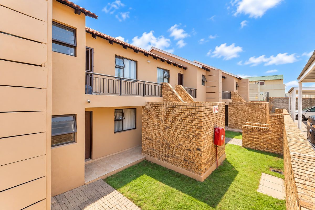 Rear view of a multi-story apartment complex featuring brick walls, a balcony with metal railings, and a small private garden area with paved walkways.