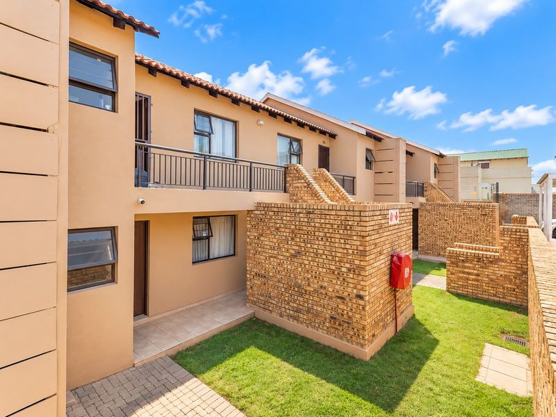Rear view of a multi-story apartment complex featuring brick walls, a balcony with metal railings, and a small private garden area with paved walkways.