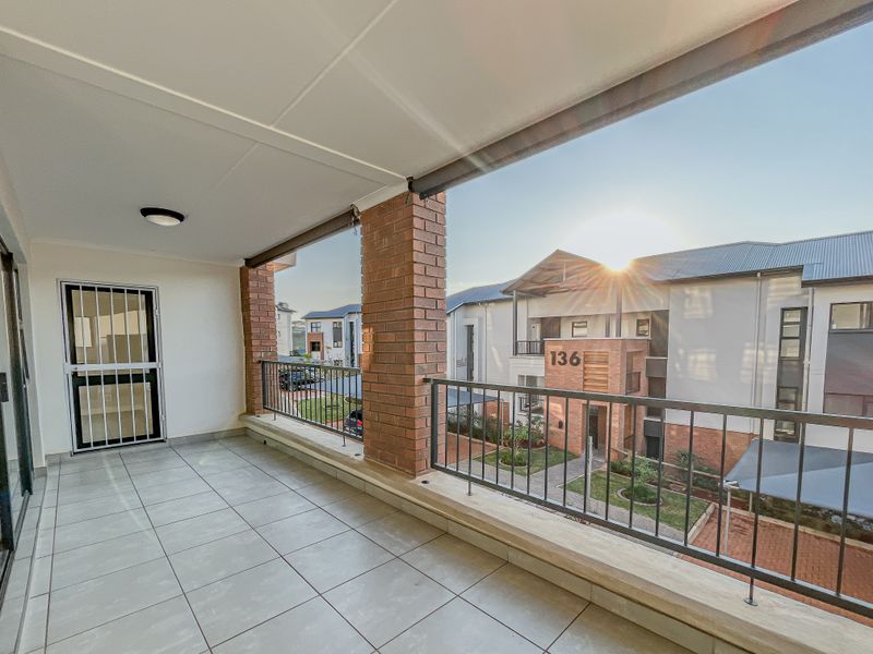 A spacious covered balcony featuring tiled flooring, brick pillars, and a metal balustrade, offering a pleasant view of the surrounding apartment complex.