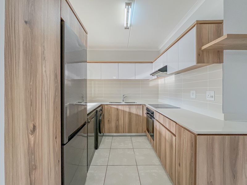 Modern galley-style kitchen featuring wood-grain cabinetry, white high-gloss upper cabinets, stone countertops, integrated appliances, and neutral tiled flooring.
