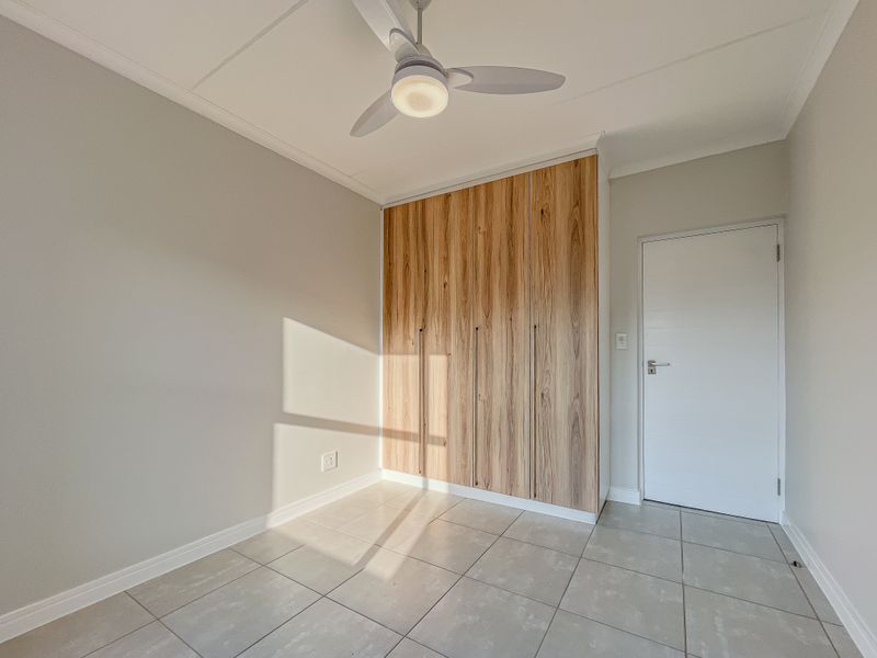 A bright, empty bedroom featuring modern wood-finish built-in cupboards, neutral tiled flooring, and a ceiling fan.