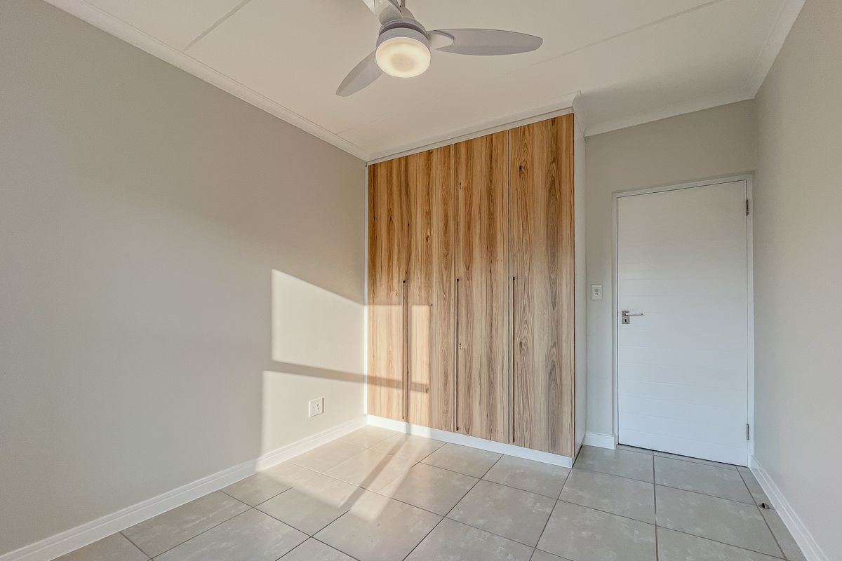 A bright, empty bedroom featuring modern wood-finish built-in cupboards, neutral tiled flooring, and a ceiling fan.