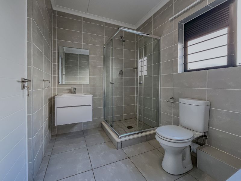 A modern, clean bathroom featuring a glass shower enclosure, wall-mounted vanity with basin, and a standard toilet, finished with neutral floor-to-ceiling wall tiles.
