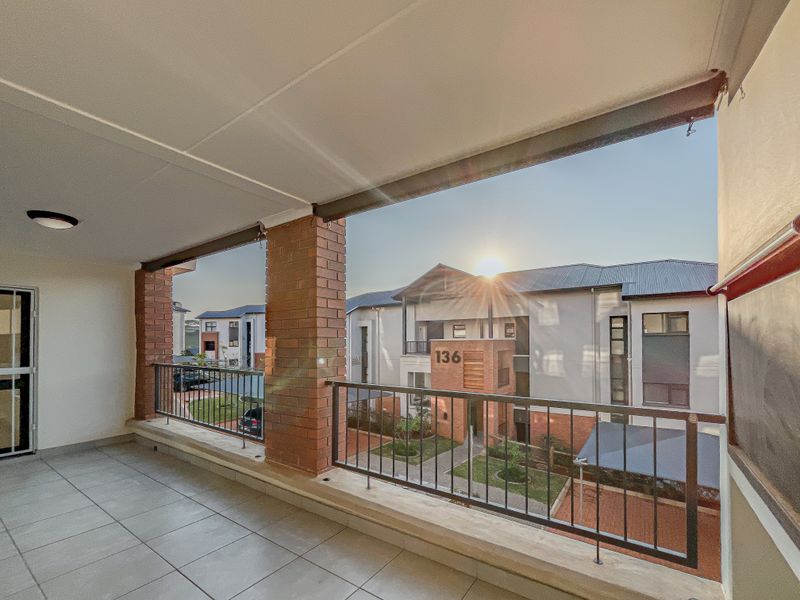 A spacious covered balcony featuring tiled flooring, brick support pillars, and a metal balustrade, overlooking the apartment complex grounds.