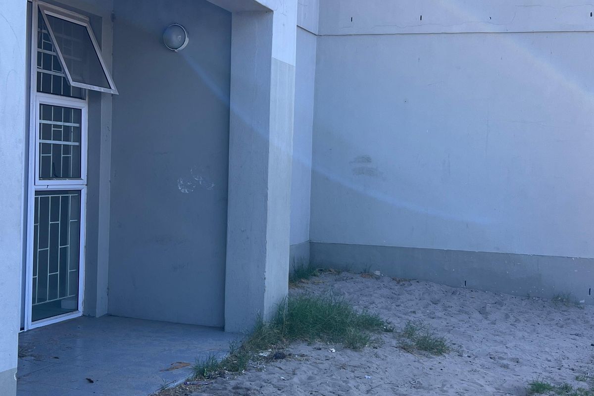 A rear exterior view of an apartment unit featuring a small balcony, security gate, and an unlandscaped sandy ground area.