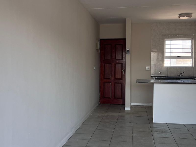 An open-plan living and kitchen area featuring neutral tiled flooring, a functional kitchen counter with sink, and a solid wood entrance door.
