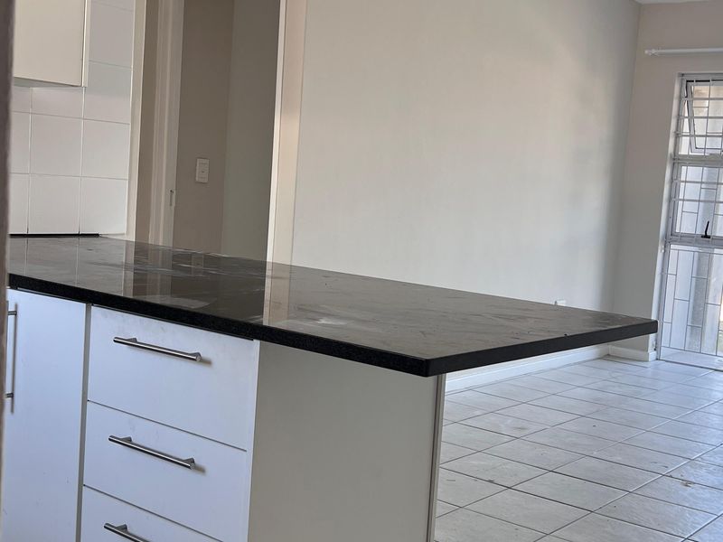 An open-plan kitchen and living area featuring white cabinetry, dark stone countertops, and neutral tiled flooring in a clean, vacant space.
