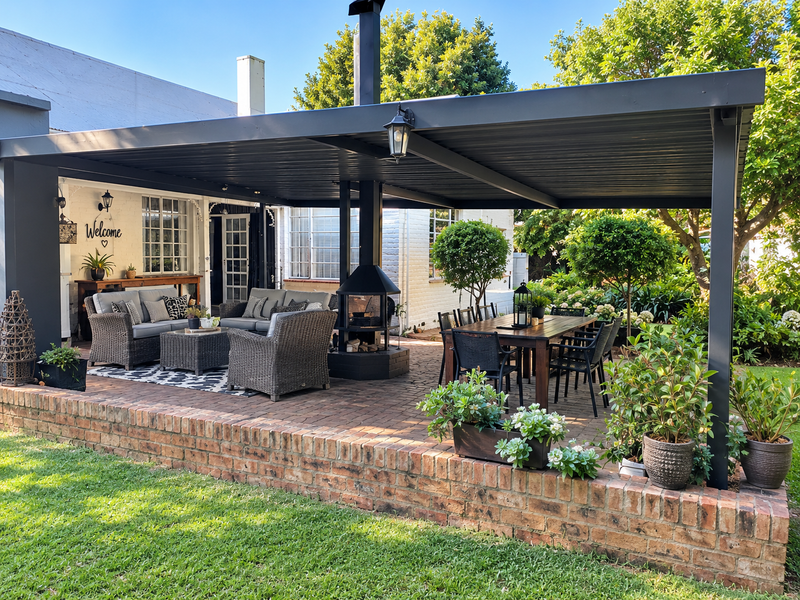 A well-maintained covered patio featuring a brick retaining wall, paved flooring, and a central freestanding fireplace, ideal for outdoor entertaining.