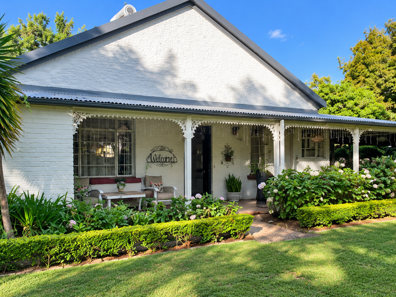 A charming white painted brick house featuring a covered veranda with decorative ironwork, surrounded by a well-maintained garden and lush landscaping.