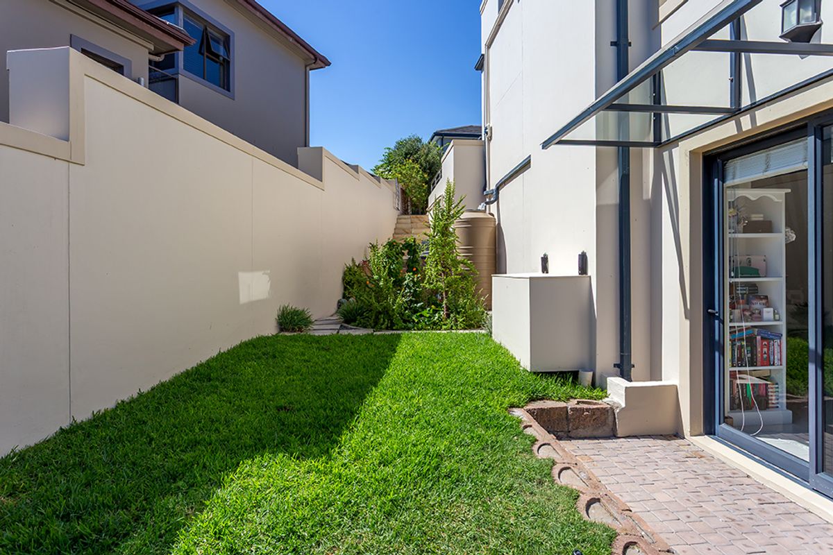 A well-maintained rear garden area featuring a paved walkway, boundary wall, water tank, and a glass awning over the sliding door entrance.