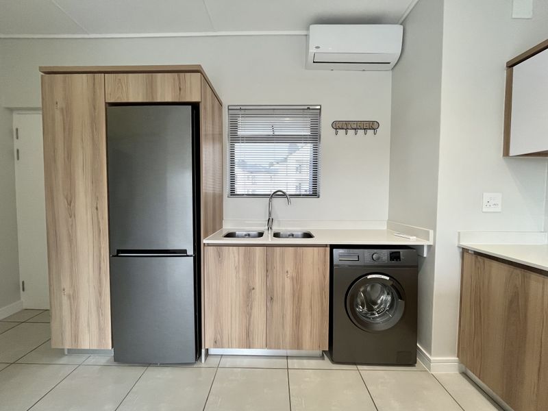 Modern kitchen area featuring wood-finish cabinetry, double sink, tiled flooring, and a wall-mounted air conditioning unit.