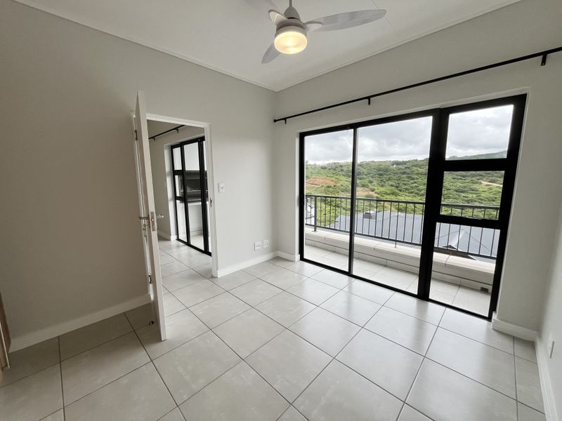 A bright, empty bedroom featuring neutral tiled flooring, a ceiling fan, and large sliding glass doors opening onto a balcony with scenic views.