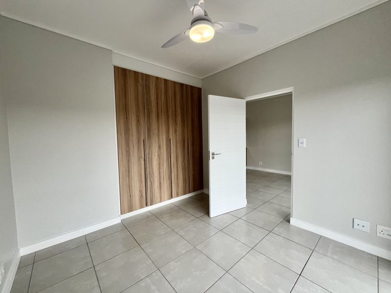 A clean, modern bedroom featuring neutral tiled flooring, a built-in wooden wardrobe, and a ceiling fan, offering a bright and versatile living space.