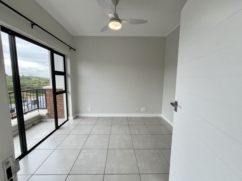 A bright, empty bedroom featuring neutral tiled flooring, a modern ceiling fan, and direct access to a private balcony through large sliding glass doors.