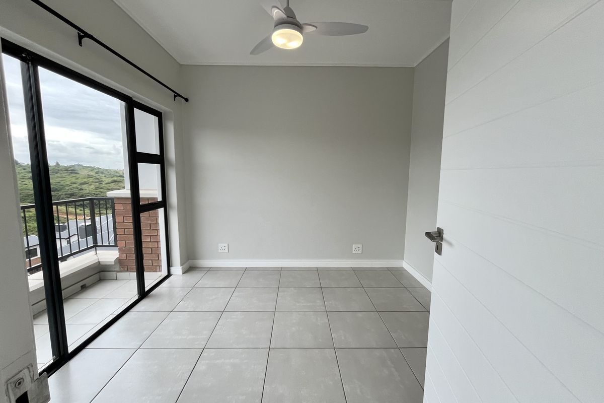 A bright, empty bedroom featuring neutral tiled flooring, a modern ceiling fan, and direct access to a private balcony through large sliding glass doors.