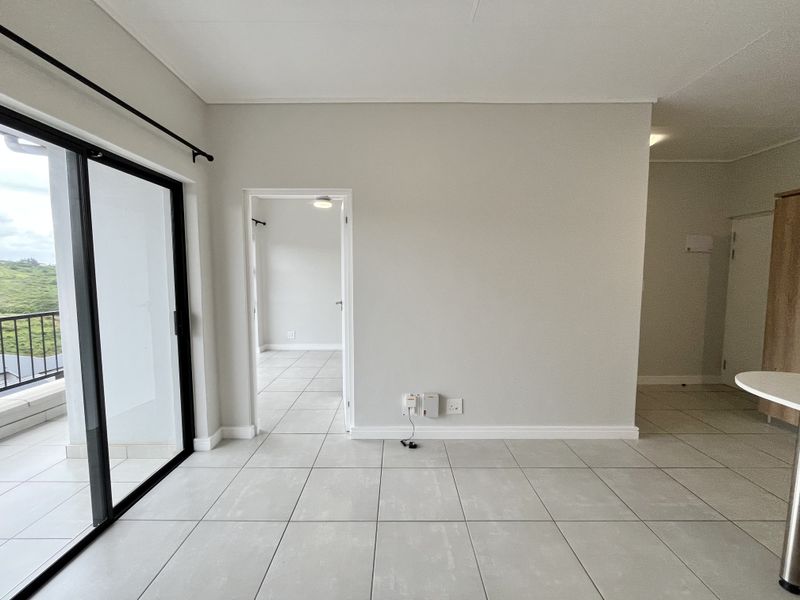 An empty, well-maintained living area featuring neutral tiled flooring, a large sliding glass door leading to a balcony, and access to an adjacent room.
