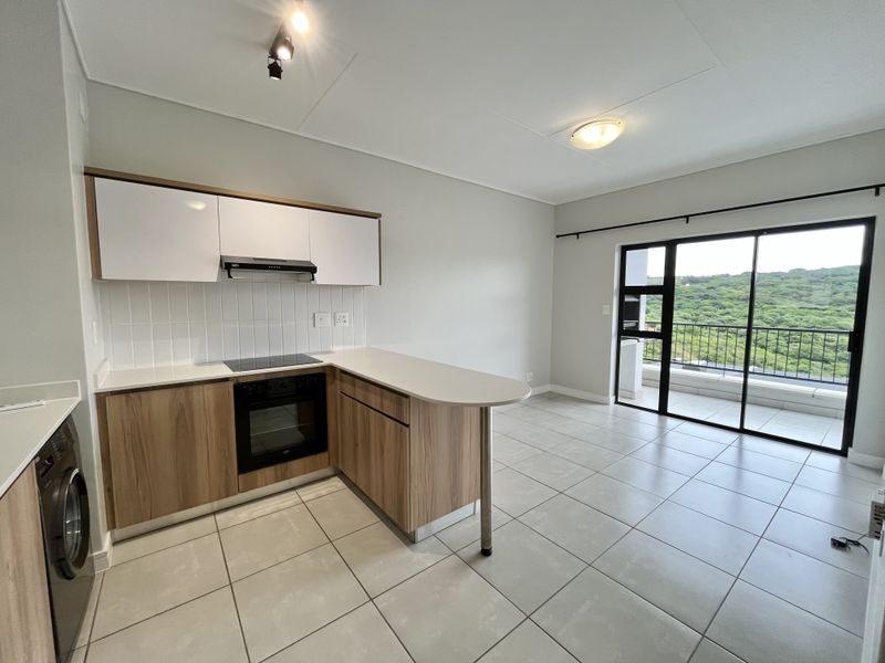 Modern open-plan kitchen featuring wood-finish cabinetry, integrated appliances, and tiled flooring, leading directly to a balcony with scenic views.