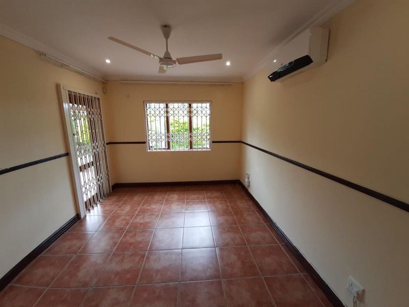 An empty living room featuring tiled flooring, a ceiling fan, a wall-mounted air conditioner, and security fixtures on the door and window.