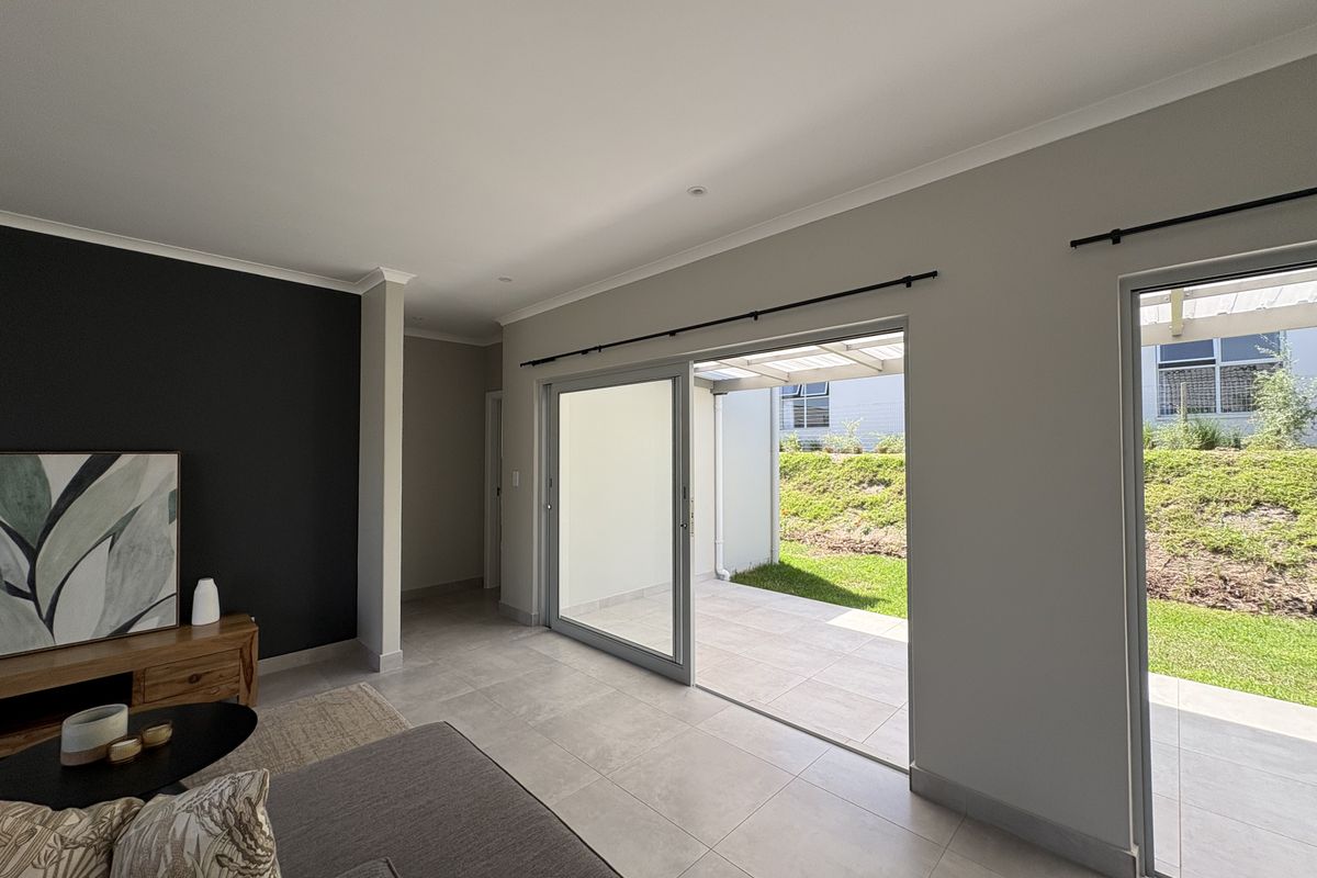 Bright living room featuring large sliding glass doors that open onto a paved patio and garden area, with neutral tiled flooring throughout.