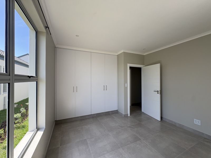 A bright, modern bedroom featuring neutral wall tones, large format floor tiles, and ample built-in cupboard storage with a view of the garden.