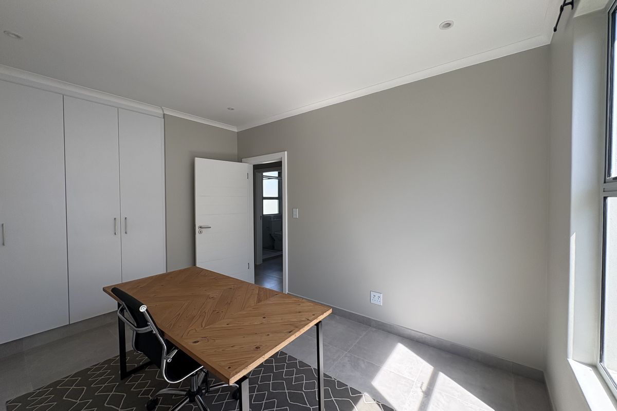 A bright bedroom featuring neutral walls, tiled flooring, built-in cupboards, and recessed lighting, with an adjoining doorway leading to an en-suite bathroom.