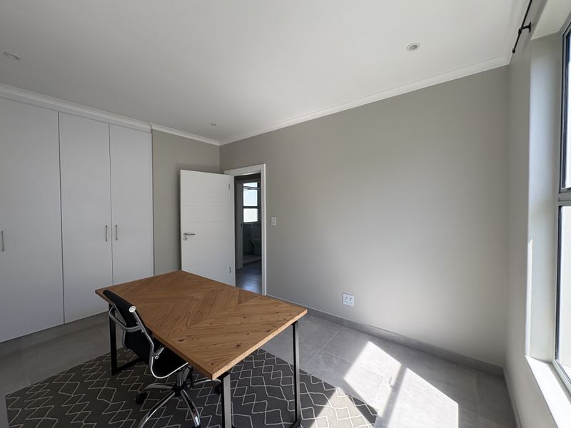 A bright bedroom featuring neutral walls, tiled flooring, built-in cupboards, and recessed lighting, with an adjoining doorway leading to an en-suite bathroom.