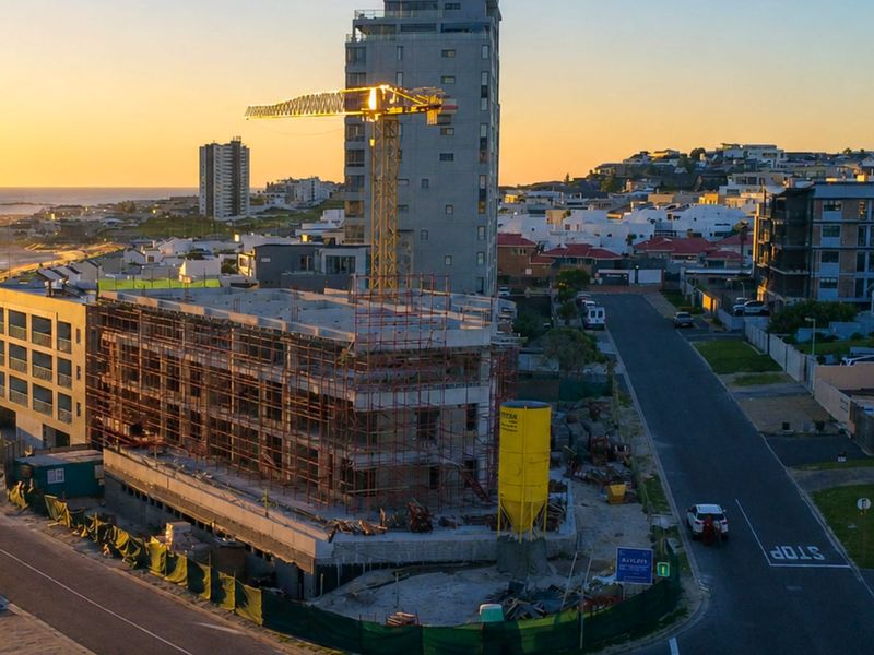 Aerial view of a multi-story apartment building under construction, featuring scaffolding and a crane, situated in a coastal neighborhood.
